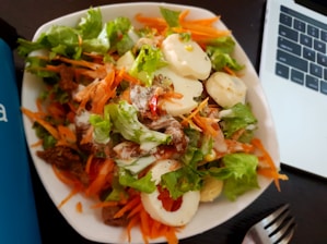 A bowl of fresh salad with leafy greens, sliced carrots, hard-boiled eggs, and roasted meat, drizzled with a creamy dressing. A laptop and a metal fork are visible nearby on a dark surface.