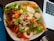 A bowl of fresh salad with leafy greens, sliced carrots, hard-boiled eggs, and roasted meat, drizzled with a creamy dressing. A laptop and a metal fork are visible nearby on a dark surface.