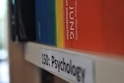 A close-up view of a bookshelf showing a psychology section label with books stored horizontally. One prominent book with a colorful spine that transitions from blue to red is partially visible.