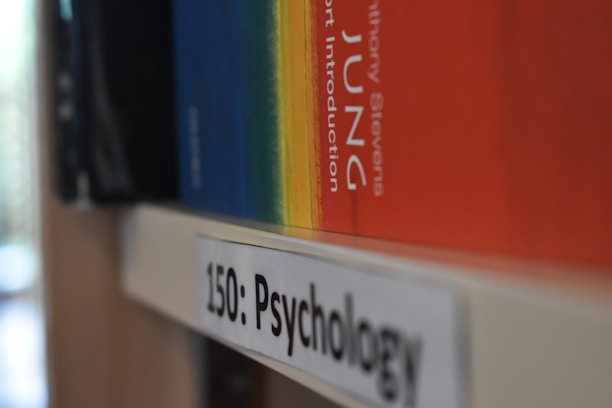 A close-up view of a bookshelf showing a psychology section label with books stored horizontally. One prominent book with a colorful spine that transitions from blue to red is partially visible.