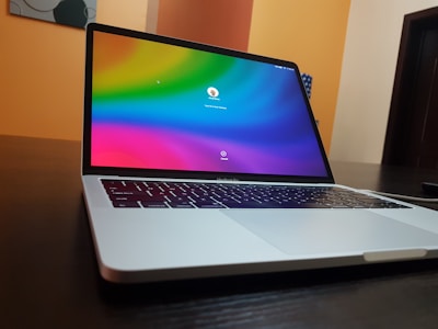 A sleek laptop sits on a dark wooden table, displaying a colorful login screen with a gradient of colors including blue, green, and red. The background includes a wall with shades of orange and cream, and part of a modern art piece.