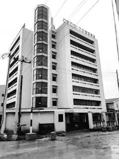 A multi-story office building with tall vertical windows and a prominent sign reading 'CEDDI TOWERS' at the top. The facade is flat with a central column of glass windows, flanked by solid walls. The environment includes power lines and a wet pavement, suggesting recent rain.