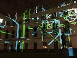 The image displays a building facade at night with a projection of abstract patterns consisting of green, blue, and white lines. There is a statue visible in the foreground, silhouetted by the light display. The windows of the building are dark, adding contrast to the bright, geometric projections.