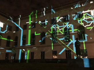 The image displays a building facade at night with a projection of abstract patterns consisting of green, blue, and white lines. There is a statue visible in the foreground, silhouetted by the light display. The windows of the building are dark, adding contrast to the bright, geometric projections.