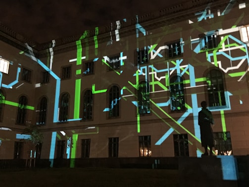 The image displays a building facade at night with a projection of abstract patterns consisting of green, blue, and white lines. There is a statue visible in the foreground, silhouetted by the light display. The windows of the building are dark, adding contrast to the bright, geometric projections.