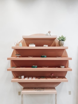 A wall-mounted shelving unit with five horizontal shelves is lined with various small decorative items and potted plants. The shelves are a natural wood color, placed against a circular backdrop. On the shelves, there are intricate pieces of jewelry, small glass vases, and artistic elements arranged in a minimalist style. The setting is calm and visually appealing, emphasizing simplicity and elegance.