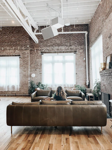 A cozy living room with exposed brick walls and warm hardwood floors, bathed in soft evening light from large industrial windows overlooking a close-up city street.
