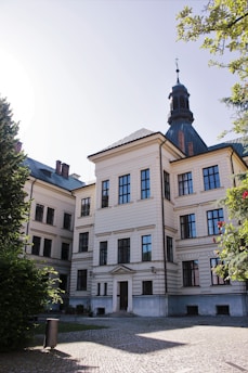 A large, classic building with white walls and numerous windows, featuring a central spire on the roof. It is surrounded by green trees and a cobblestone courtyard, with sunlight casting shadows across the ground.