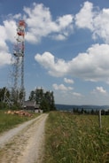 A tall communication tower stands amidst a rural landscape, surrounded by lush greenery and a gravel path leading past a small building. The sky is bright and partly cloudy, with fluffy white clouds scattered across a vivid blue background.