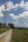 A tall communication tower stands amidst a rural landscape, surrounded by lush greenery and a gravel path leading past a small building. The sky is bright and partly cloudy, with fluffy white clouds scattered across a vivid blue background.