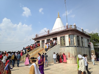 A bustling scene around a temple structure with people ascending and descending the stairs. The temple features a white, pointed dome and is adorned with gold accents. Numerous visitors, dressed in colorful attire, gather outside on a sunny day with a clear blue sky and a few clouds.