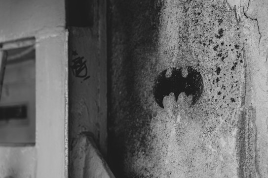 A black and white image featuring a rough textured wall with a spray-painted Batman logo in the center. The image captures an urban and gritty atmosphere with shadowy surroundings and worn surfaces.