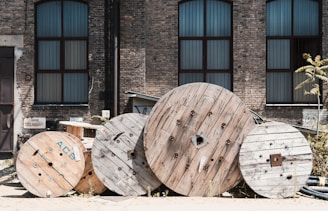Large wooden cable reels are stacked against the backdrop of an old industrial brick building with tall windows. The windows are framed with dark metal, and the bricks are weathered, giving a rustic feel. Some weeds and a small tree are growing between the reels, adding a touch of greenery.