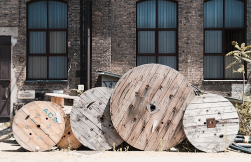 Large wooden cable reels are stacked against the backdrop of an old industrial brick building with tall windows. The windows are framed with dark metal, and the bricks are weathered, giving a rustic feel. Some weeds and a small tree are growing between the reels, adding a touch of greenery.