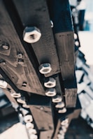 A close-up view of a wooden structure with numerous metal hexagonal nuts attached, creating an industrial and mechanical feel. The nuts are evenly spaced and aligned along the dark wooden beams.