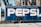 A large delivery truck features the bold branding of a well-known soft drink company with its name prominently displayed in large white letters on a blue background. The truck is parked next to an old brick building with tall, industrial-style windows. Shadows from overhead cables stretch across the scene, adding an urban feel.