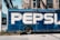A large delivery truck features the bold branding of a well-known soft drink company with its name prominently displayed in large white letters on a blue background. The truck is parked next to an old brick building with tall, industrial-style windows. Shadows from overhead cables stretch across the scene, adding an urban feel.