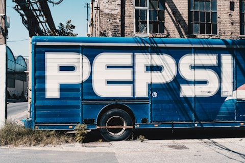 A large delivery truck features the bold branding of a well-known soft drink company with its name prominently displayed in large white letters on a blue background. The truck is parked next to an old brick building with tall, industrial-style windows. Shadows from overhead cables stretch across the scene, adding an urban feel.
