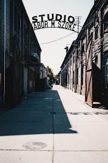 An industrial alleyway with large iron gates stands open, casting shadows on the sunlit pavement. The central view focuses on the overhead sign reading 'STUDIO' in large letters. The architecture is mostly brick with visible wires and piping along the walls.