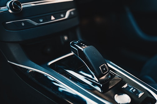 The interior of a car reveals a modern gear shift surrounded by a glossy black finish. Additional controls and buttons are visible, with sleek metallic accents enhancing the sophisticated design.