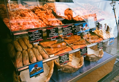 A display case filled with various seafood items such as prawns, crab claws, squid, and fish fillets. In front of the seafood are baskets of baguettes. Small signs with descriptions are placed in front of each food item.