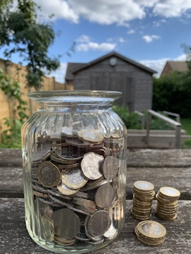 A glass jar filled with various coins is placed on a weathered wooden surface. Next to the jar are stacks of coins. In the background, there is a garden with a wooden shed, greenery, and a clear blue sky with scattered clouds.