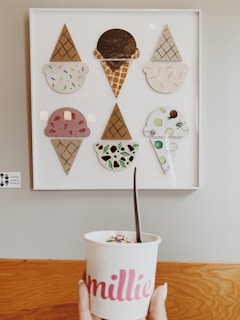 A wall-mounted art piece displays various stylized ice cream designs, each with different flavors and toppings, arranged in a white frame. Below the artwork, a hand is holding a cup labeled 'millie,' filled with ice cream and topped with colorful sprinkles. The background consists of a pale wall and wooden surface.