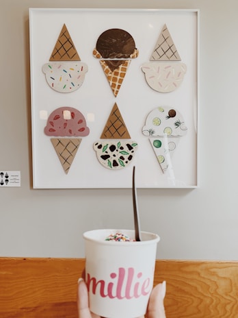 A wall-mounted art piece displays various stylized ice cream designs, each with different flavors and toppings, arranged in a white frame. Below the artwork, a hand is holding a cup labeled 'millie,' filled with ice cream and topped with colorful sprinkles. The background consists of a pale wall and wooden surface.