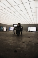 A dimly lit room with a grid-patterned illuminated ceiling creates a futuristic atmosphere. Several screens display close-up images of eyes. In the foreground, a professional camera is positioned on the floor, focusing towards the screens.