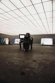 A dimly lit room with a grid-patterned illuminated ceiling creates a futuristic atmosphere. Several screens display close-up images of eyes. In the foreground, a professional camera is positioned on the floor, focusing towards the screens.