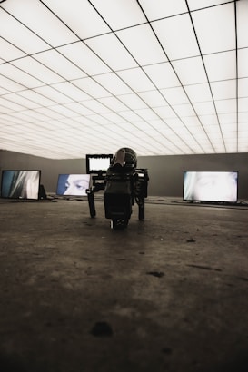 A dimly lit room with a grid-patterned illuminated ceiling creates a futuristic atmosphere. Several screens display close-up images of eyes. In the foreground, a professional camera is positioned on the floor, focusing towards the screens.