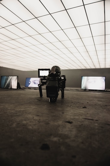 A dimly lit room with a grid-patterned illuminated ceiling creates a futuristic atmosphere. Several screens display close-up images of eyes. In the foreground, a professional camera is positioned on the floor, focusing towards the screens.