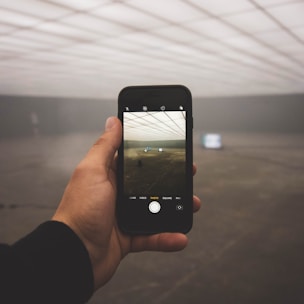 A hand holds a smartphone, capturing a photograph of a large, empty, indoor space. The ceiling is designed with grid-like lighting, and the floor is bare, with fog obstructing visibility in the background. The phone displays the same scene being photographed.