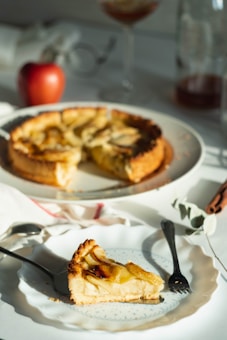 A slice of apple pie is placed on a white plate with a fork beside it. The rest of the pie, missing one slice, sits on a broader plate in the background. Sunlight casts a warm glow on the scene, highlighting the golden crust and apple slices. An apple and some decorative elements, like eucalyptus and cinnamon sticks, accompany the setup.