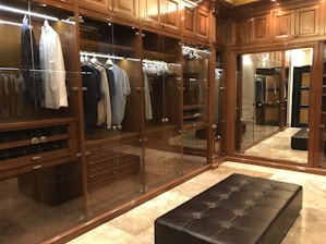 A luxurious walk-in closet features wooden cabinetry with glass doors. Inside, a variety of shirts and jackets are neatly hung on hangers, illuminated by subtle lighting. The room has polished stone flooring and a plush leather bench in the center, reflecting an elegant and organized space.