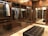 A luxurious walk-in closet features wooden cabinetry with glass doors. Inside, a variety of shirts and jackets are neatly hung on hangers, illuminated by subtle lighting. The room has polished stone flooring and a plush leather bench in the center, reflecting an elegant and organized space.
