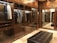 A luxurious walk-in closet features wooden cabinetry with glass doors. Inside, a variety of shirts and jackets are neatly hung on hangers, illuminated by subtle lighting. The room has polished stone flooring and a plush leather bench in the center, reflecting an elegant and organized space.