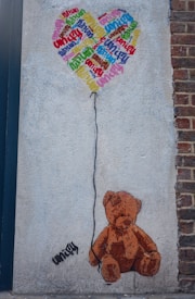 A teddy bear is painted on a wall, sitting at the bottom with a balloon string leading up to a heart-shaped balloon. The balloon is composed of the word 'unity' repeatedly in various colors and orientations. The wall has a rough texture, and there's a brick surface visible on the side.