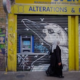 A person walks past a storefront with rolled-down shutters featuring graffiti art. An ATM is accessible on the left side, surrounded by colorful graffiti on a yellow and brick wall. The shutters display a black and white bird illustration. The person is wearing a headscarf and long dark clothing.