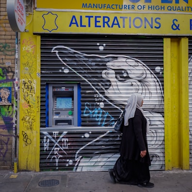 A person walks past a storefront with rolled-down shutters featuring graffiti art. An ATM is accessible on the left side, surrounded by colorful graffiti on a yellow and brick wall. The shutters display a black and white bird illustration. The person is wearing a headscarf and long dark clothing.