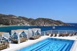 A luxurious seaside view featuring a swimming pool with intricate tile patterns. Surrounding the pool are several cabanas adorned with white drapes and sun loungers made of wood. Beyond the pool, a deep blue sea stretches out to meet rugged, rocky hills. A sailboat is visible on the water, and a small beach with buildings is nestled along the shore in the distance.