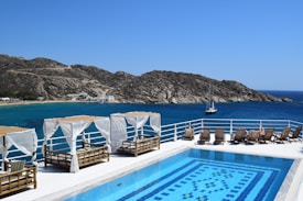 A luxurious seaside view featuring a swimming pool with intricate tile patterns. Surrounding the pool are several cabanas adorned with white drapes and sun loungers made of wood. Beyond the pool, a deep blue sea stretches out to meet rugged, rocky hills. A sailboat is visible on the water, and a small beach with buildings is nestled along the shore in the distance.