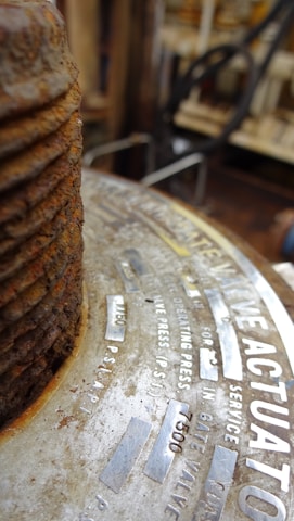 Close-up view of a metal surface with text engraved on it. The focus is on a rusty, corroded, industrial component. The text includes technical specifications, possibly related to pressure or mechanical components.