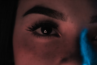 A close-up of a human eye with a triangle reflection in the pupil. The eyebrow is well-defined, and there's a blue light effect partially visible on the right side of the image.
