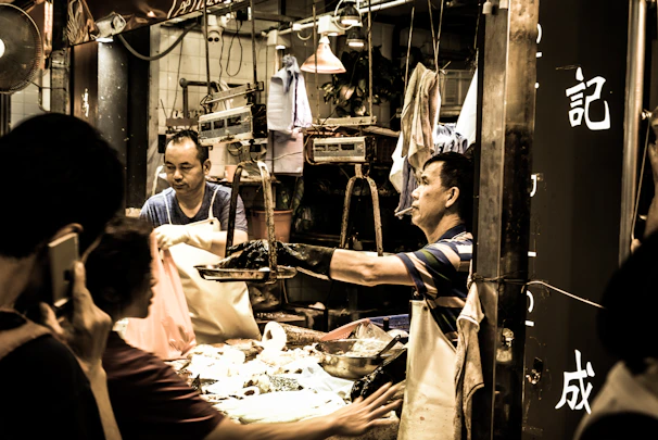 Friendly vendor handing a customer a bag of goods with a smile in a busy neighborhood market setting.