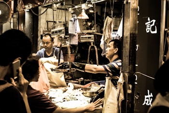 A busy street market scene where vendors are interacting with customers. One vendor wearing an apron is handing over something to a customer while another is seen in the background handling goods. The setting is lively and energetic with various market items and scales visible around.