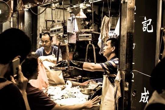 A busy street market scene where vendors are interacting with customers. One vendor wearing an apron is handing over something to a customer while another is seen in the background handling goods. The setting is lively and energetic with various market items and scales visible around.