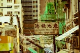 A cityscape with a real estate sign in the foreground, prominently displaying the name and phone number. Surrounding the sign are various buildings with different architectural styles, some with scaffolding and green netting. The scene features a mix of older, worn structures alongside modern glass-fronted buildings. Power lines and construction materials add to the urban complexity.