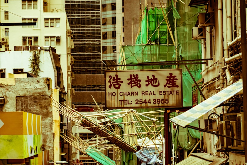 A cityscape with a real estate sign in the foreground, prominently displaying the name and phone number. Surrounding the sign are various buildings with different architectural styles, some with scaffolding and green netting. The scene features a mix of older, worn structures alongside modern glass-fronted buildings. Power lines and construction materials add to the urban complexity.