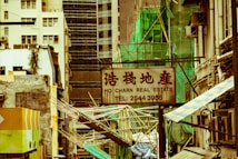 A cityscape with a real estate sign in the foreground, prominently displaying the name and phone number. Surrounding the sign are various buildings with different architectural styles, some with scaffolding and green netting. The scene features a mix of older, worn structures alongside modern glass-fronted buildings. Power lines and construction materials add to the urban complexity.